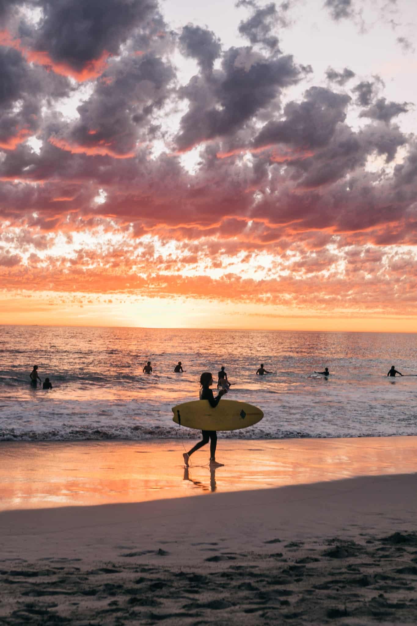 Unrecognizable people in swimwear surfing and swimming on sandy summer beach in calm sea at sundown