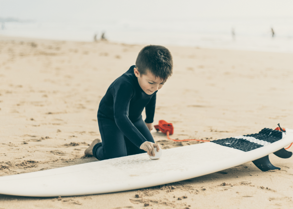 young boy applying surfboard wax to his surfboard