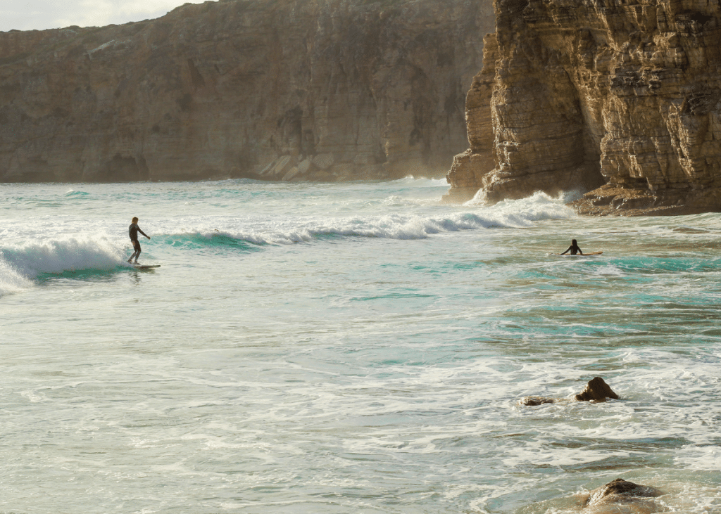 Two surfers catch waves near a rugged cliff coastline under a warm, ambient light during the best tide for surfing. The ocean is lively and clear blue, creating a serene and adventurous setting.
