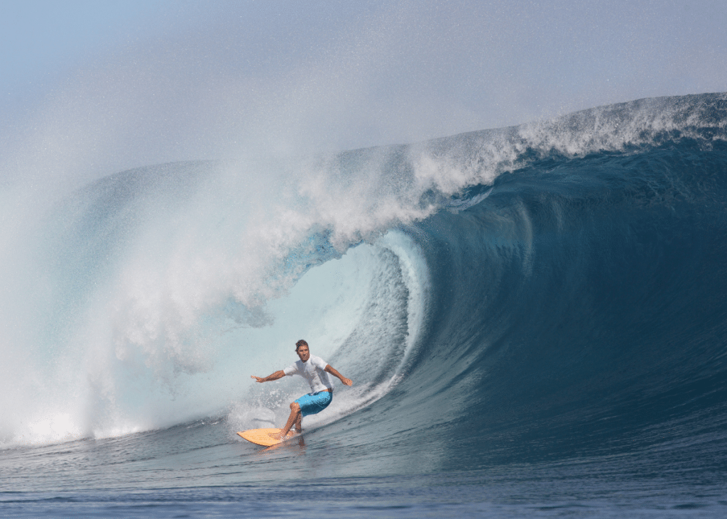 A surfer expertly rides a large, curling blue wave at the best tide for surfing in the ocean, posing with arms outstretched for balance on a bright orange surfboard, with clear