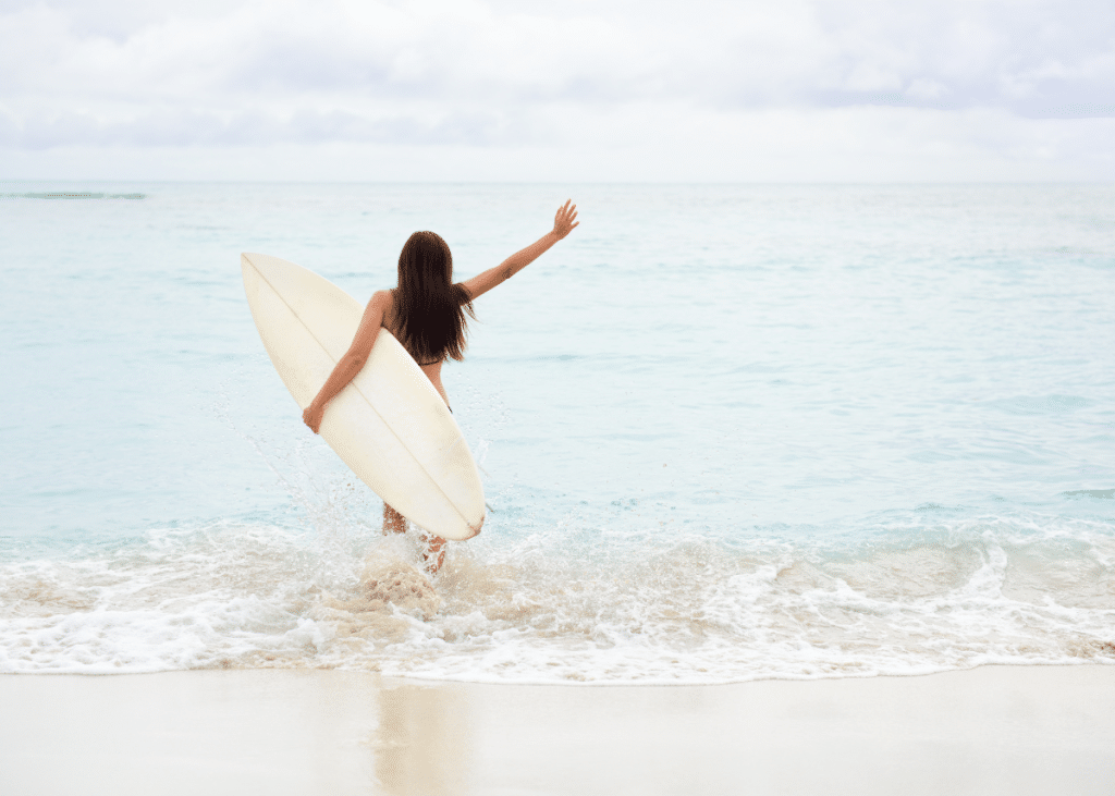 A woman with long dark hair, holding a surfboard, runs into the ocean waves, raising one arm in excitement under a cloudy sky.