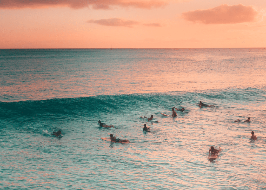 Surfers practicing surf etiquette in an ocean with gentle waves at sunset, under a soft-hued sky, cast in a warm pink and orange glow.