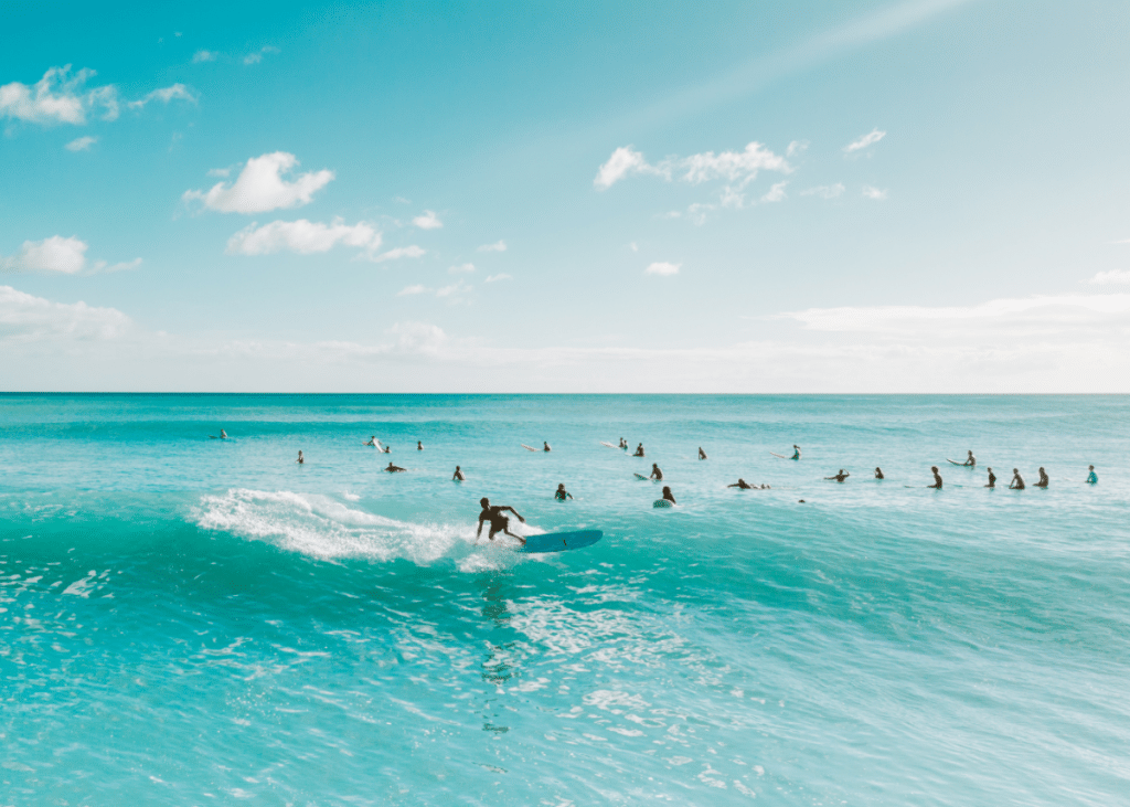 A group of surfers practicing surf etiquette in the ocean, with one person adeptly riding a wave in the foreground. The clear blue sky and turquoise sea create a serene backdrop.