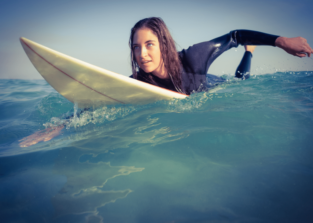 A woman in a wetsuit chosen for surfing paddles a surfboard through the clear blue water, looking intently to the side.