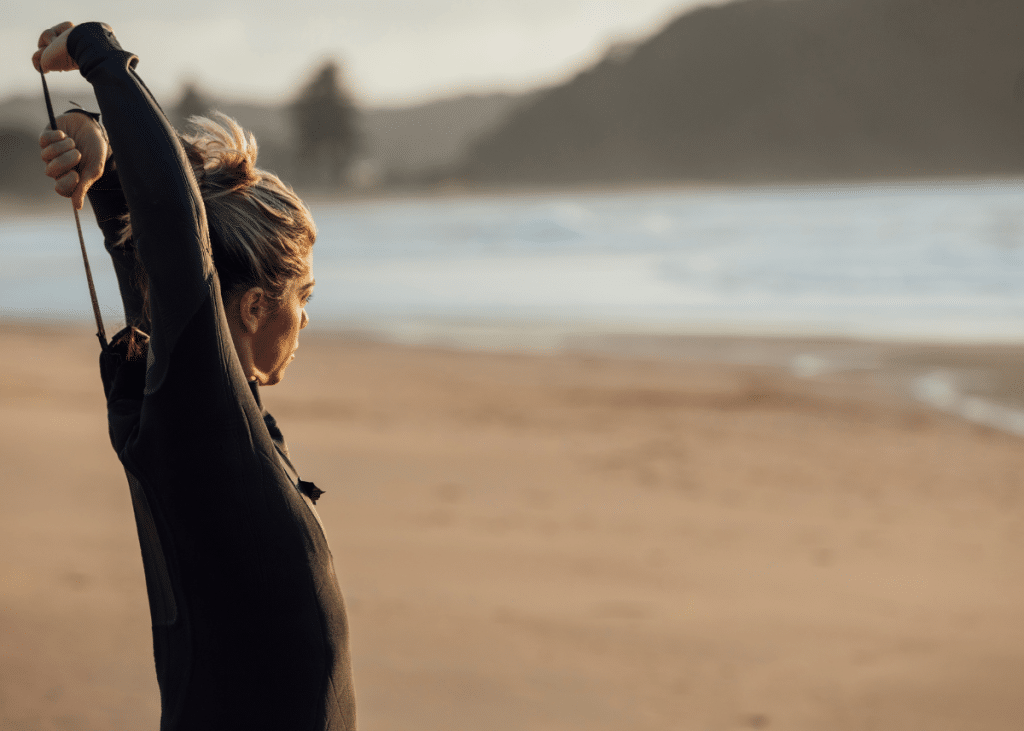 A woman stretching on a beach at sunset, wearing a wetsuit for surfing, with ocean waves and cliffs in the background.