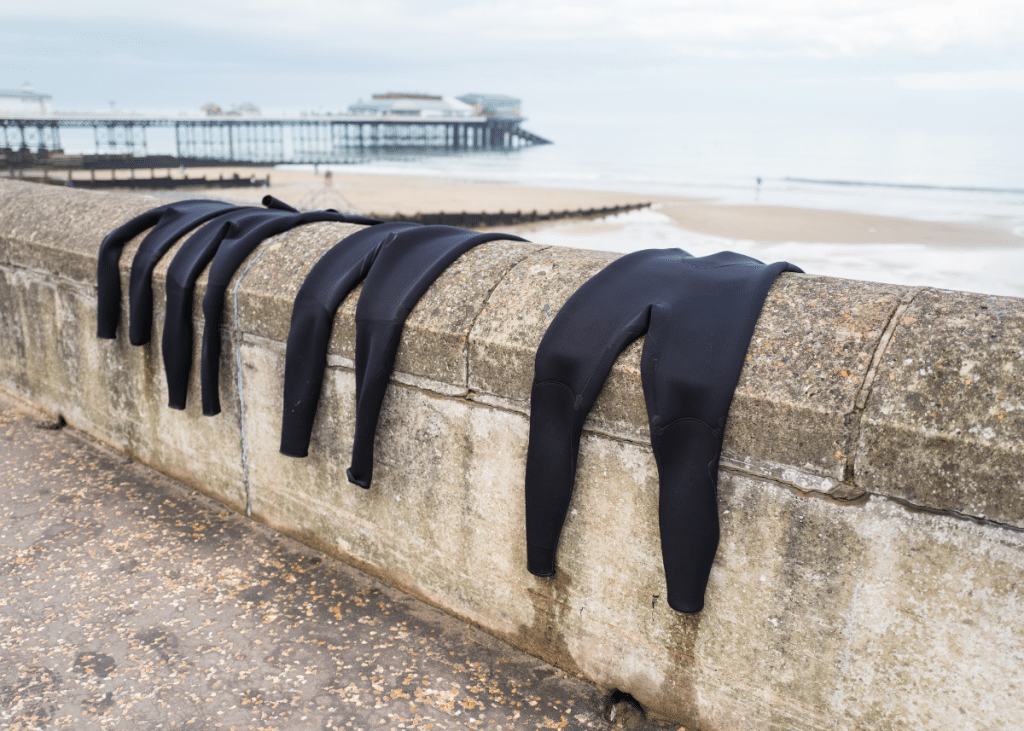 Wetsuits designed for surfing draped over a seaside wall to dry, with a sandy beach and a pier in the background under a cloudy sky.