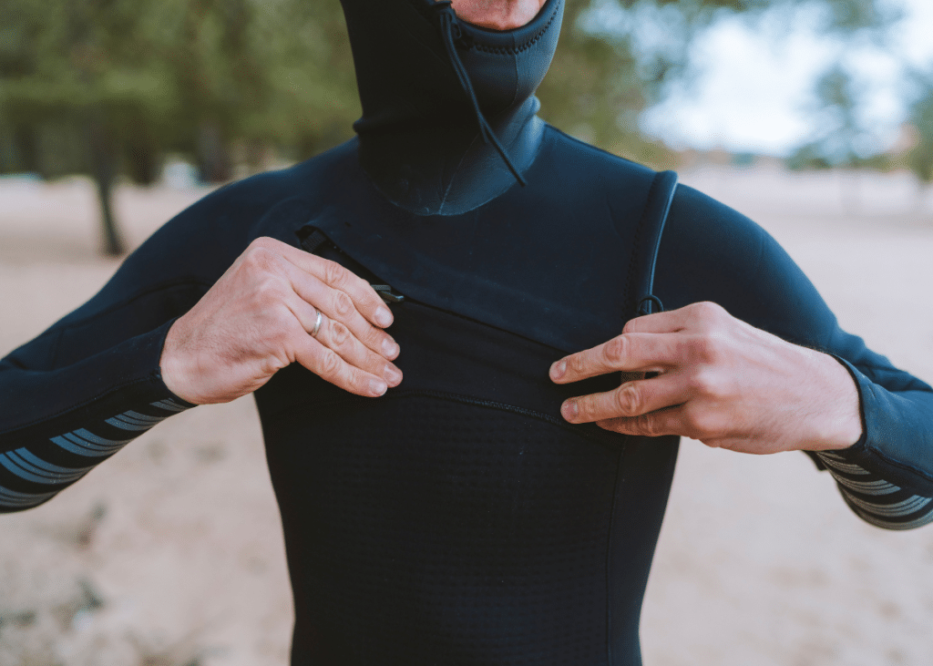 A person in a black wetsuit for surfing adjusts the collar near the beach, focusing on the zipper area with a sandy background visible.
