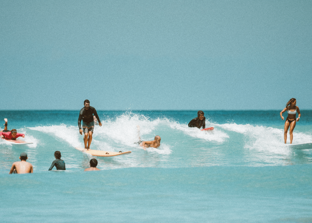 People engaging in various water activities in the ocean. Some are surfing on waves, including a few kooks, as onlookers watch from the water. The scene is captured in a vibrant, summ
