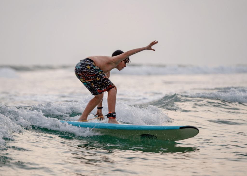 A young surfer, often called a kook, balancing on a blue surfboard, riding a gentle wave in the ocean. He's wearing brightly patterned board shorts and appears focused on maintaining his