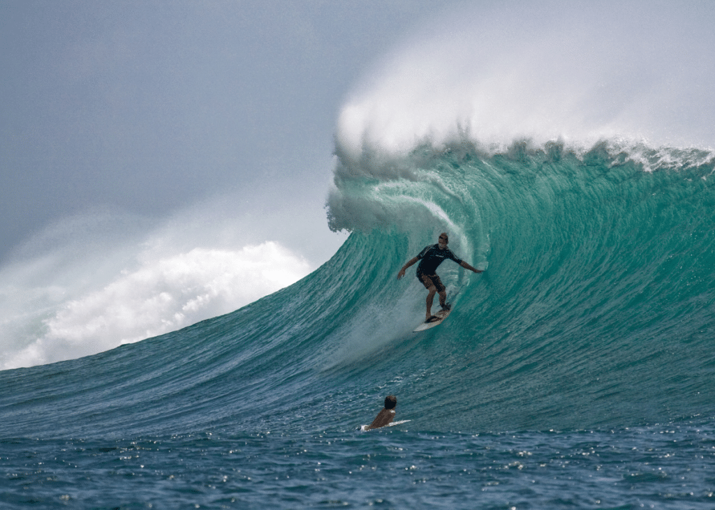 A surfer rides a large, powerful wave, demonstrating skill and balance, while another kook watches from the water below under a cloudy sky.
