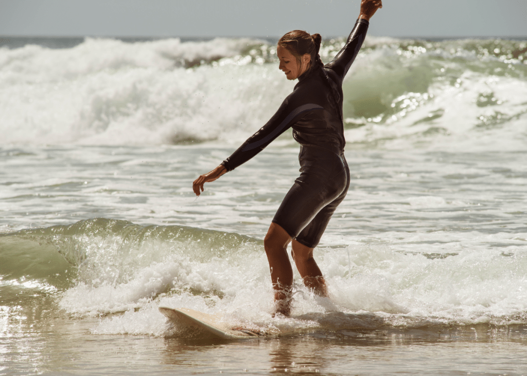 A beginner woman in a wetsuit skillfully surfing a small wave at the beach, with sunlight highlighting the texture of the water and her focused expression.