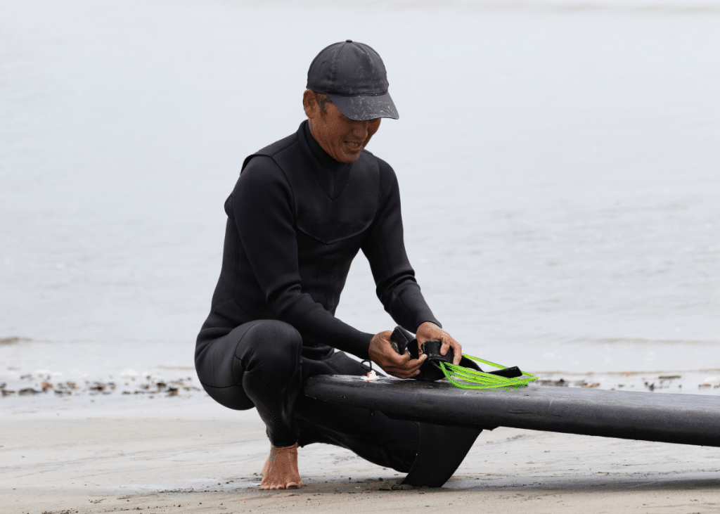 A beginner in a wetsuit crouches by a black surfboard, attaching green fins, on a sandy beach.