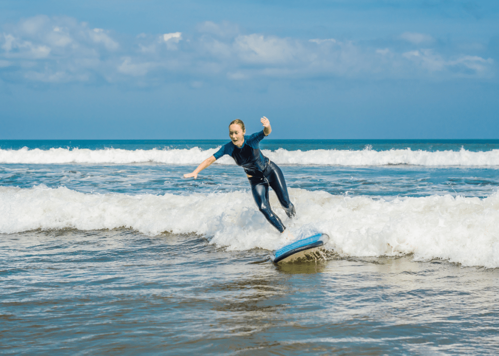 A young beginner boy in a wetsuit surfs a small wave, balancing on a blue surfboard with a bright sky and ocean in the background. His expression is focused and joyful.