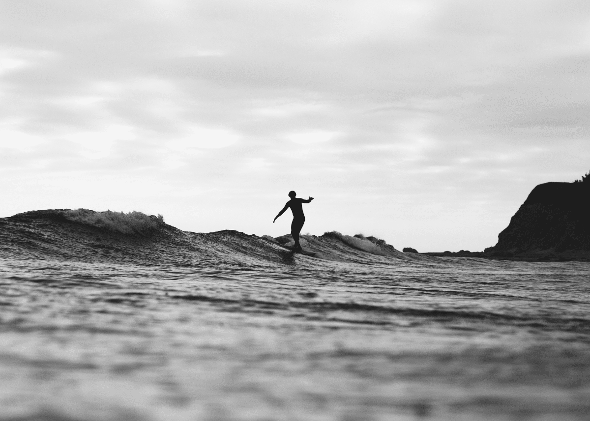 A silhouette of a beginner surfer riding a wave at a beach, captured in black and white, with a rugged coastline visible in the background.