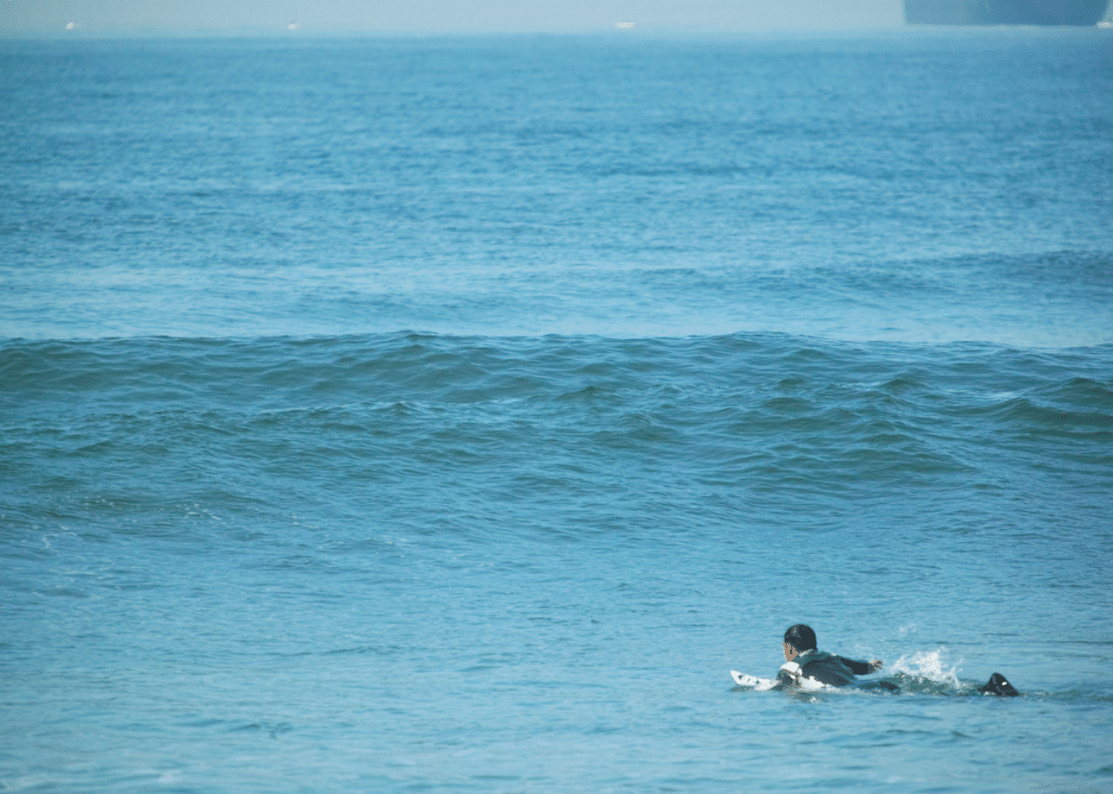A beginner surfer in a wetsuit paddling on a calm blue sea under clear skies.
