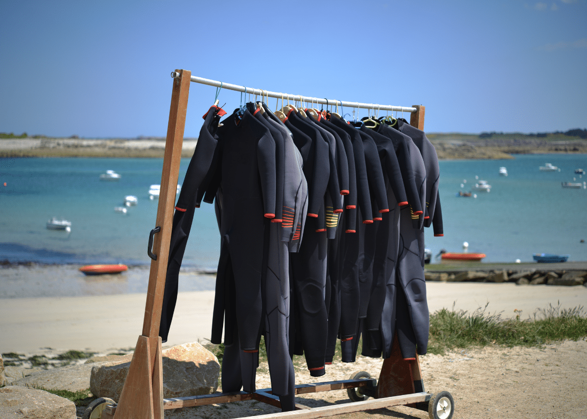 A rack of black wetsuits with red and yellow trim, ideal for surfing, hanging outdoors, with a scenic backdrop of a calm blue sea dotted with small boats and a clear sky.